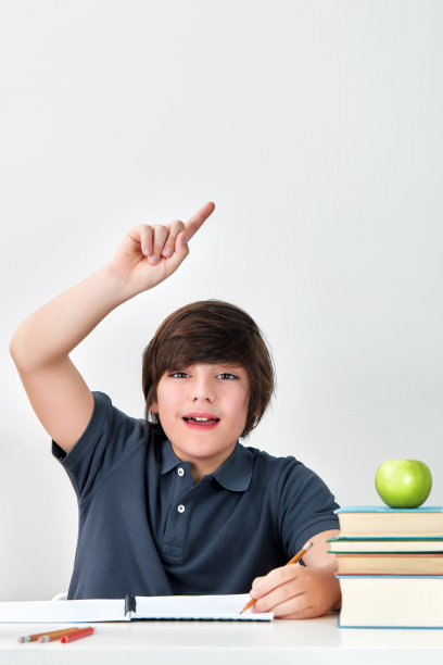 在学校教室里，兴奋而快乐的白人男孩坐在课桌旁用食指举起手回答问题图片下载