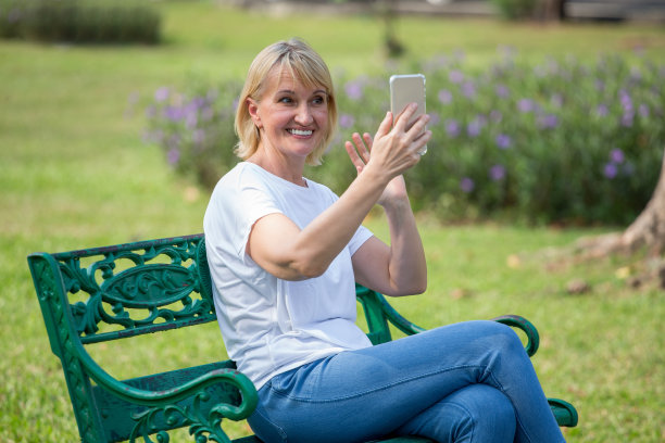 快乐的老女人放松在公园使用智能手机自拍照片。老年人坐在夏季公园看手机和视频通话。老年女性休息。成熟的关系图片下载