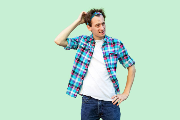 肖像的深思熟虑的年轻人在休闲蓝色格子衬衫和头巾站着，抓着他的头和思考该做什么。图片下载