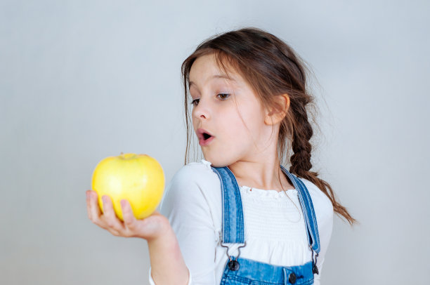 情感肖像小美丽的女孩扎着辫子在牛仔裤工作服吃一口拿着苹果。6 - 7年的工作室图片下载