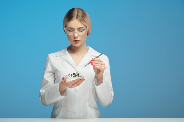 年轻漂亮的金发女人穿着白色外套，戴着护目镜，背景是蓝色。拿着镊子和一片叶子。科学的概念，对植物和产品的研究。转基因生物测试。科学家，生物学家，化学家，微生物学家。图片下载