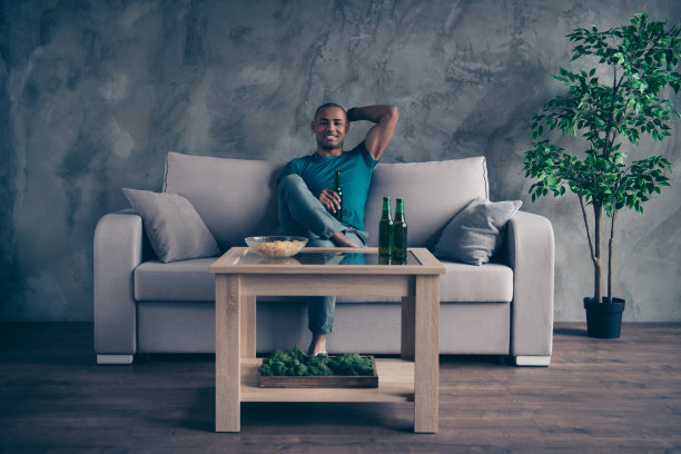 全身的身体尺寸照片惊人的他他他的深色皮肤男子帅气的手绿色瓶子啤酒苹果酒土豆油炸快餐暂停休息穿蓝色的t恤裤子坐在舒适的沙发房间房子在室内图片下载