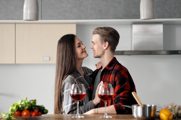 一对穿着格子衬衫的漂亮年轻夫妇在厨房里准备食物，用玻璃杯喝红酒。图片下载