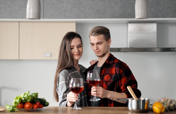 一对穿着格子衬衫的漂亮年轻夫妇在厨房里准备食物，用玻璃杯喝红酒。图片下载