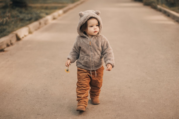 兜帽里的可爱宝宝手里拿着一颗黄色的蒲公英，在路上走着。图片下载