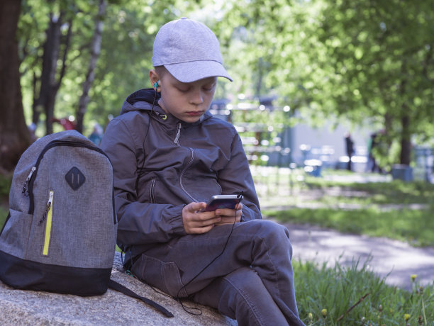 孩子拿着手机坐在公园里的一块大石头上。男孩看着屏幕，使用应用程序，玩游戏。阳光明媚的一天。人、科技、休闲理念图片下载