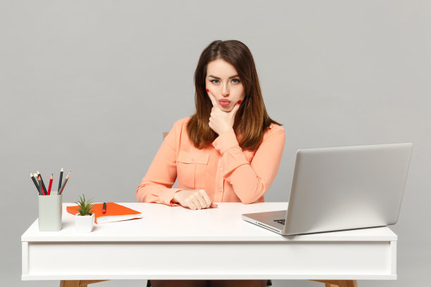 年轻迷人的女人在柔和的休闲衣服把手撑在下巴上坐在桌子上工作与电脑笔记本电脑隔离在灰色的背景。成就商业事业生活理念。模拟拷贝空间。图片下载