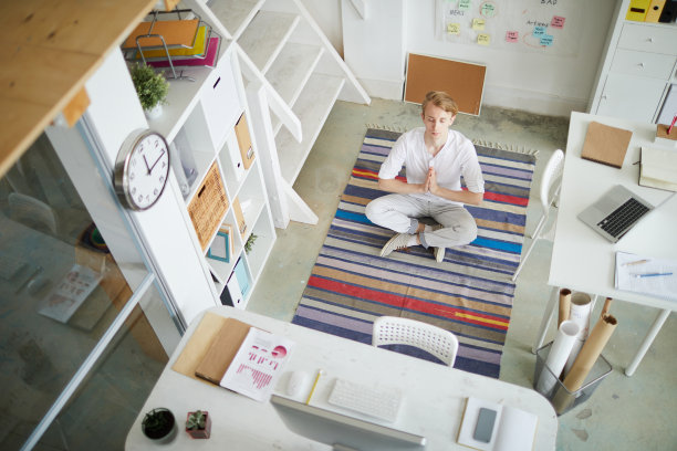 高角度冷静集中的年轻男性经理在白色衣服坐在交叉双腿和双手合十礼，而冥想在舒适的办公室图片下载