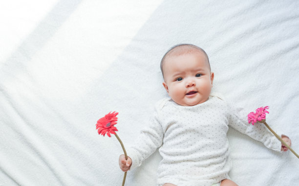 亚洲女婴拿着红粉相间的花朵，白色的毛巾背景和阳光阴影。小女孩正在玩红色和粉红色的花。宝宝对她的花很满意。图片下载