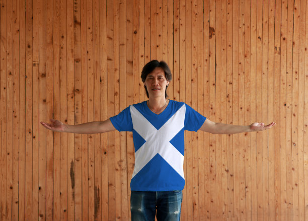 男子穿着苏格兰国旗颜色的衬衫，双臂张开站在木墙的背景上。图片下载