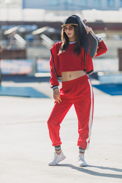 年轻女性的肖像戴着黑帽子，红色衣服滑板手拿着她的滑板。带着滑板在户外看摄像机的女人图片下载