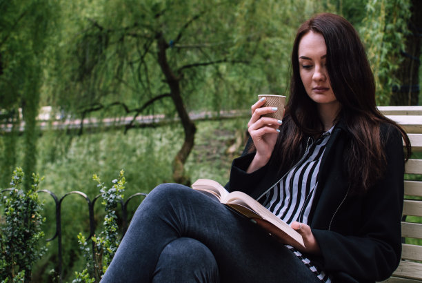 沉思的黑发女孩坐在公园的长椅上，喝着纸杯咖啡，读着一本软皮书。图片下载