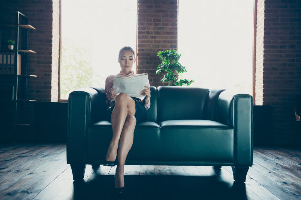 画像漂亮迷人优雅时尚自信内容别致的女士鲨鱼合格专家坐在沙发上阅读小报在现代工业阁楼室内风格的工作场所工作站图片下载