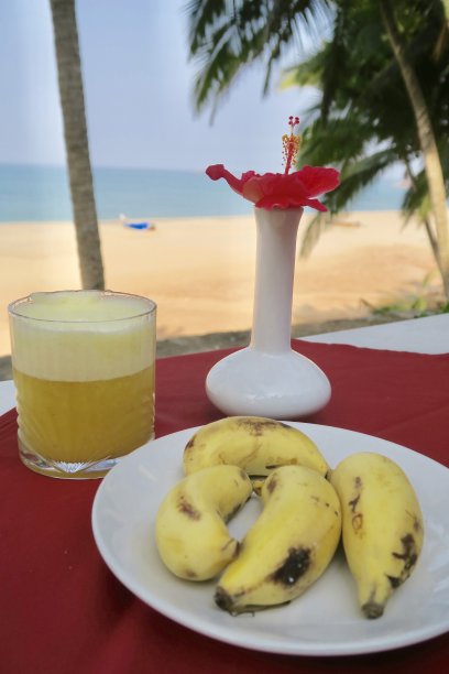 简单的热带早餐的形象，新鲜的菠萝果汁，小印度香蕉，红色的木槿花花瓶，喀拉拉邦的海滩背景，印度南部，鲜花安排早餐餐桌和户外的阳光，喝果汁图片下载