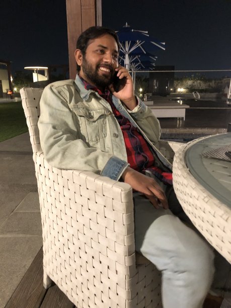 英俊的印度男子使用手机，坐在外面的咖啡桌在晚上捂着耳朵讲电话，笑着和微笑的手机照片，白色的柳条桌子和椅子庭院设置，玻璃顶部和黑色的夜空，家具图片下载