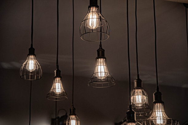 天花板上的金属灯罩用黑色缆绳吊着。玻璃灯泡内有闪亮的灯丝。各种钢丝骨架形状的几何形状。带有深色背景的闪亮吊灯。图片下载
