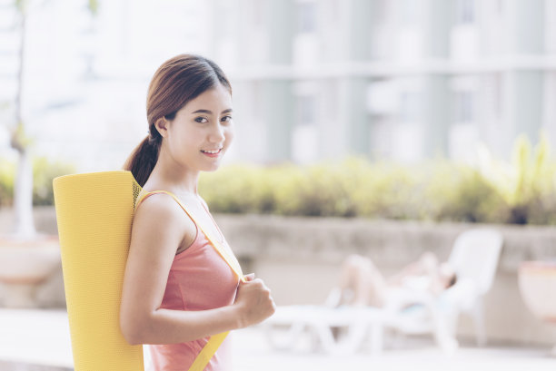 健康美丽的女人拿着瑜伽垫在她的肩膀看相机和微笑。医疗保健的概念图片下载