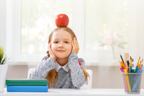 回到学校。小学生女孩坐在桌前，头上戴着一个红苹果。孩子在模糊的窗户背景下学习。教育观念。图片下载