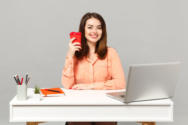 年轻的微笑女子在柔和的衣服拿着纸杯咖啡或茶坐在办公桌上工作，电脑笔记本电脑孤立在灰色的背景。成就商业事业生活理念。模拟拷贝空间。图片下载