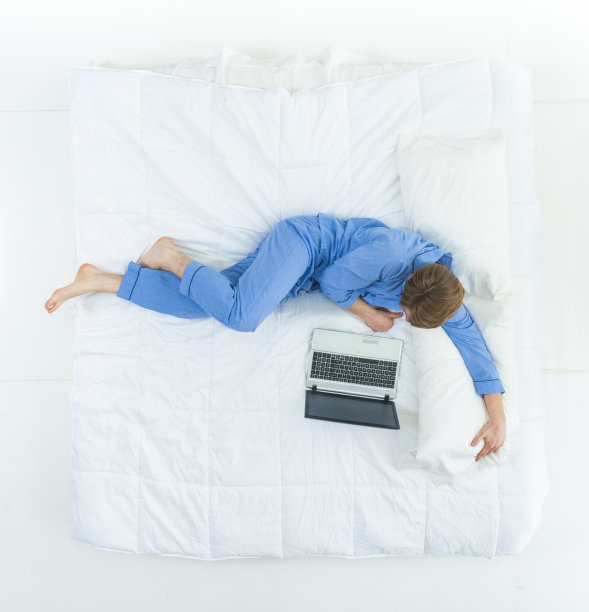 英俊的男人穿着睡衣躺在床上用着他的工作笔记本电脑图片下载