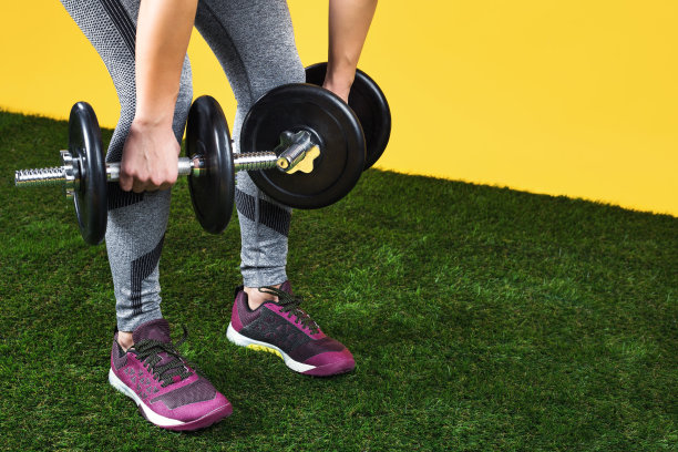 近距离的照片，英俊的年轻女子腿练习与哑铃在绿色的草地在黄色的背景。图片下载