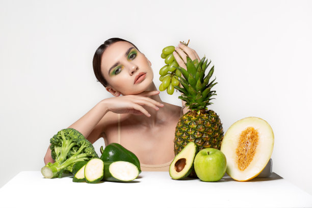 美丽的年轻女子图片与水果和蔬菜在桌子上，手持绿色葡萄孤立在灰色的背景图片下载