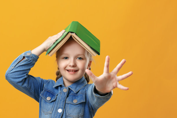 黄色背景上一个有趣的小女孩的肖像。这个学生的头上有一本书。孩子伸出手掌，击掌。图片下载