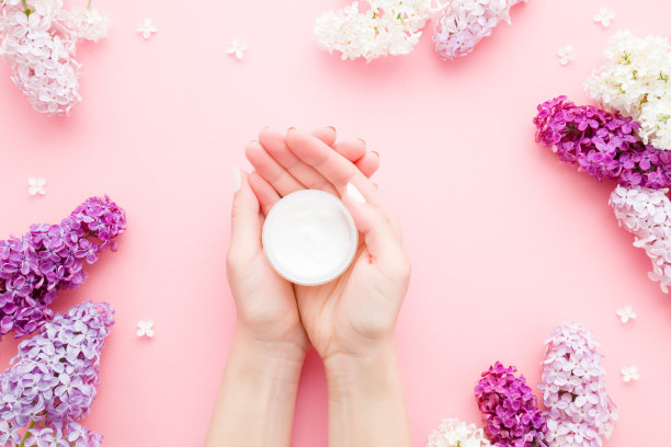 年轻，完美的女人手里拿着白色奶油罐在粉红色的桌子上。关心指甲和清洁，柔软，光滑的身体皮肤。美丽的枝条新鲜，鲜艳的丁香花。前视图。图片下载