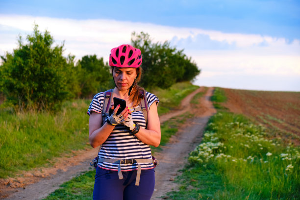日落时分，一名女子骑着自行车在乡间的MTB路线上查看手机，以一条通向地平线的土路为背景。图片下载