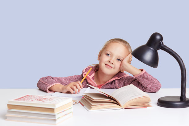微笑迷人的小女孩坐在桌子旁，和她的妈妈一起做作业，试着写作文，愉快地看，用阅读灯好视力，孤立在白色的工作室墙壁上图片下载