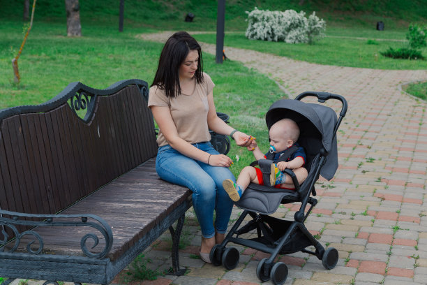 在一个阳光明媚的夏日里，美丽的母亲抱着婴儿车里的婴儿坐在长椅上图片下载