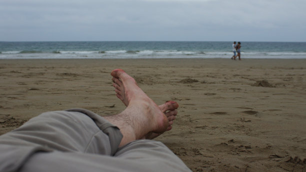 一个男人光着脚躺在沙滩上，背景是大海图片下载