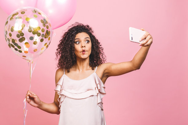 非洲裔美国积极女孩的自画像穿着气球在手拍摄自拍前相机孤立在粉红色的背景。图片下载