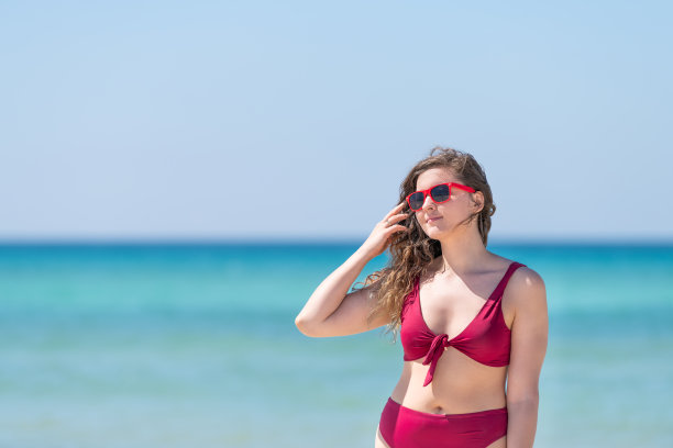 在佛罗里达圣罗莎，一名年轻的微笑女子身穿红色比基尼泳装，戴着太阳镜，用海水做背景，抚摸着头发图片下载