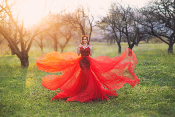 女王身着鲜亮优雅的红裙，拖着飘逸的长尾长裙摆姿势拍摄艺术照片，一头黑发、头戴皇冠的女子双手搭在肩膀上，就像森林里的女巫在清晨的阳光中像水母一样图片下载