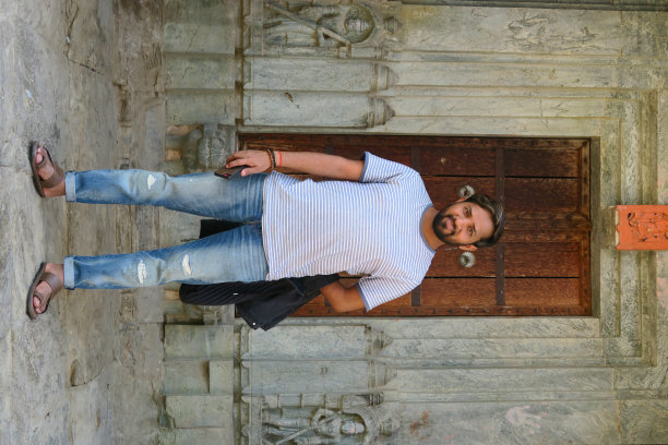 一个快乐的印度男人穿着破洞牛仔裤，蓝白相间的t恤，拿着手机和帽衫，站在木门前，脚上穿着皮凉鞋，英俊的印度男人微笑着，灰色的头发突出，性感的短胡子整洁图片下载