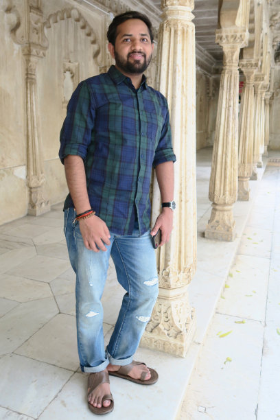 英俊的年轻印度男子游客在传统模板与拱门和圆柱，观光男子拿着手机，靠在华丽的石柱，穿着牛仔裤和蓝/绿格子衬衫，rakhi手镯和皮革凉鞋图片下载