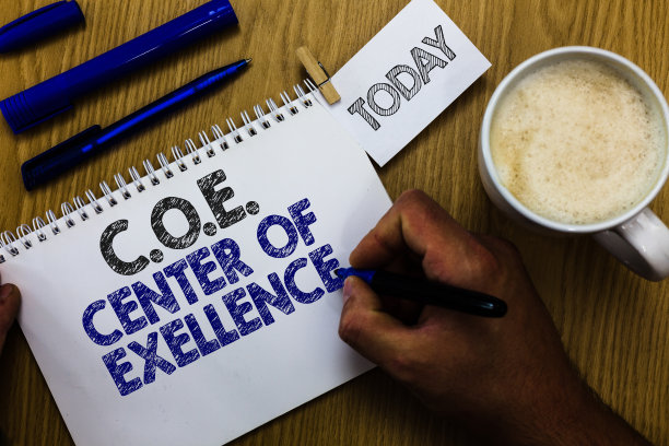 文字标志显示C.O.E卓越中心。概念照片成为阿尔法领袖在你的位置实现男人拿着马克笔笔记本衣服夹提醒木制桌子杯子咖啡。图片下载
