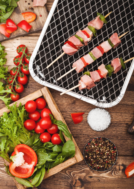 生猪肉串和辣椒粉在一次性的木炭烧烤架上，新鲜的蔬菜在木制的背景用叉子和刀。盐、胡椒粉、生菜、辣椒粉和圣女果。俯视图图片下载