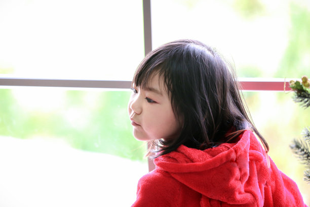 一个穿红衣服的女孩站在窗边。女孩站在窗边向旁边看。独自在家的孩子。图片下载