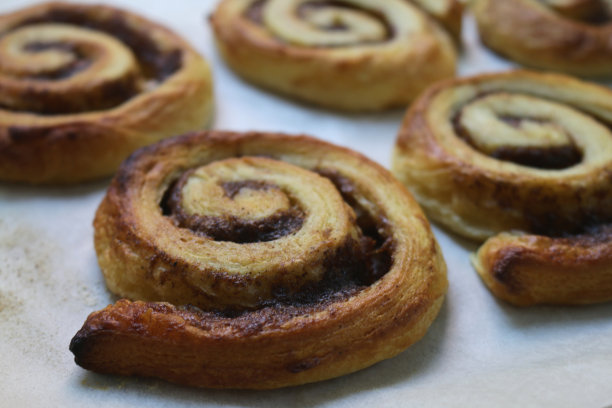 自制肉桂漩涡糕点的图片，在厨房的烤箱里用蓬松的糕点和糖霜淋烤，甜丹麦糕点肉桂漩涡食谱在防油纸烤盘上，为欧式早餐糕点，漩涡/蜗牛蛋糕图片下载