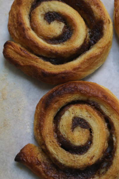 自制肉桂漩涡糕点的图片，在厨房的烤箱里用蓬松的糕点和糖霜淋烤，甜丹麦糕点肉桂漩涡食谱在防油纸烤盘上，为欧式早餐糕点，漩涡/蜗牛蛋糕图片下载