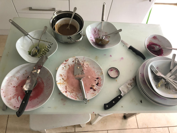 家庭晚餐后，白色的长方形玻璃餐桌上摆放着肮脏的盘子、刀叉和平底锅，厨房水池里的凳子等着清洗。凌乱的厨房需要用白色的橱柜和碗柜进行清理和整理图片下载