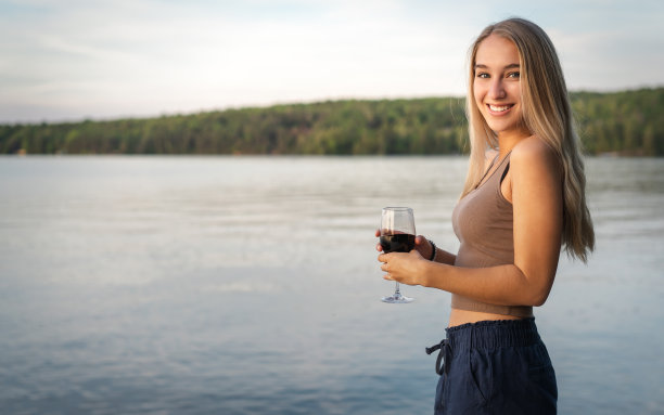 美丽的年轻女子在夏天和她的一杯红酒在一个平静的湖在日落图片下载