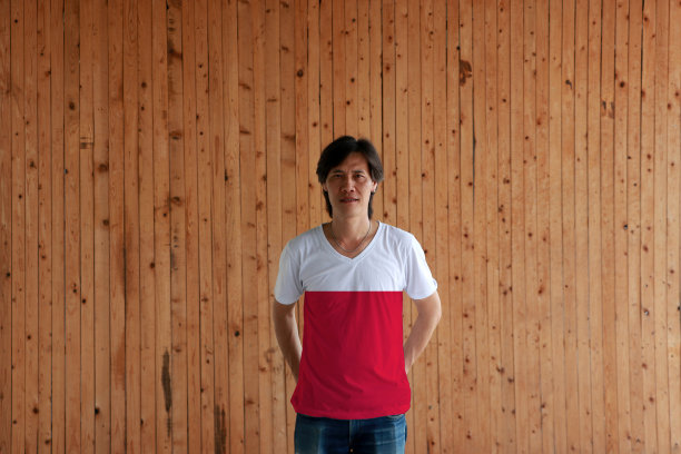 一名男子身穿波兰国旗色衬衫，双手交叉站在背后的木墙背景上。图片下载