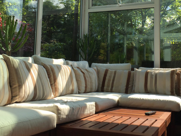 白色UPVC温室玻璃房间的图像有大窗户/ l形的角落沙发，奶油色、米色和棕色条纹软垫/枕头在温室家具上，柚木板条咖啡桌和电视电视遥控器，花园景观图片下载