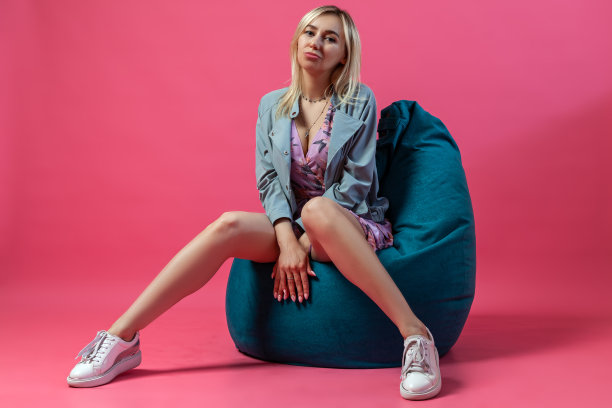 美丽的金发女孩穿着蓝色的夹克坐在一个绿色的包椅上，她的双腿分开成三角形，双手交叉在一起，在一个粉红色的孤立背景上图片下载