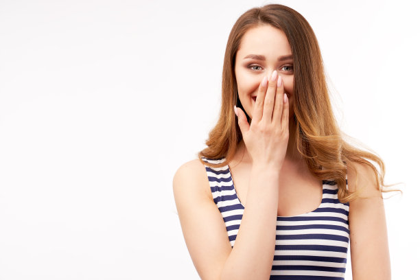 开朗愉快的可爱女性，用手捂着嘴，正面微笑，穿着休闲条纹衣服，孤立在工作室的白色墙壁上图片下载