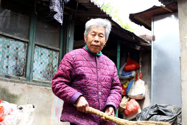 北京胡同里，一位年长的中国妇女拿着扫帚微笑着站在她的院子里。图片下载