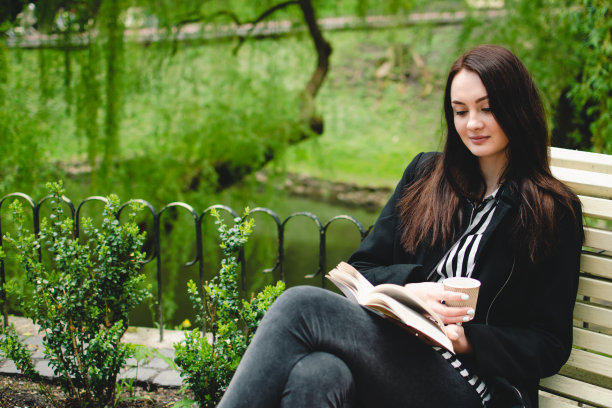 微笑的黑发女孩坐在公园的长椅上读一本软皮书。图片下载